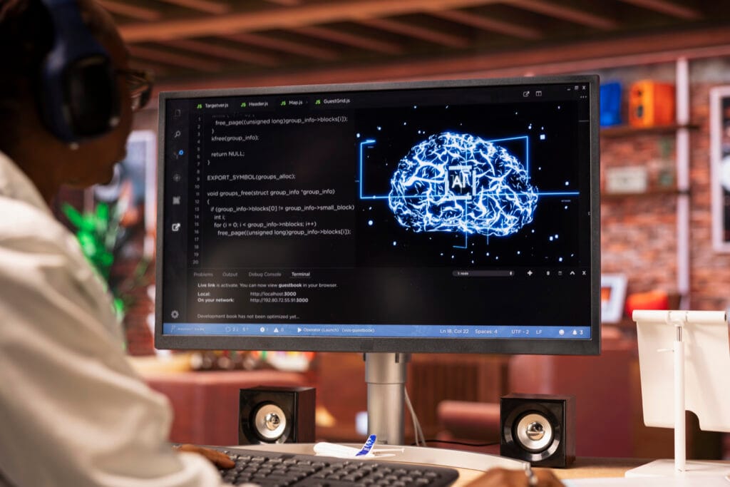 Person wearing headphones at a desk, coding on a monitor that shows code on the left and a glowing blue brain graphic on the right.