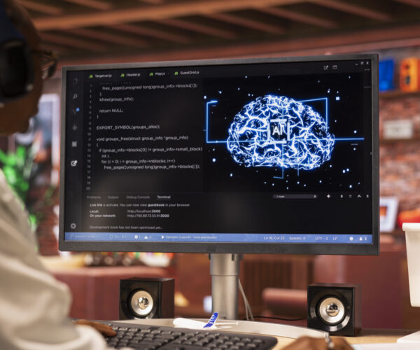 Person wearing headphones at a desk, coding on a monitor that shows code on the left and a glowing blue brain graphic on the right.