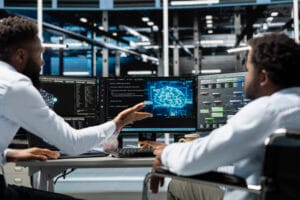 Two engineers collaborate at a workstation, monitoring three screens showing code and a holographic brain graphic in a tech office setting.