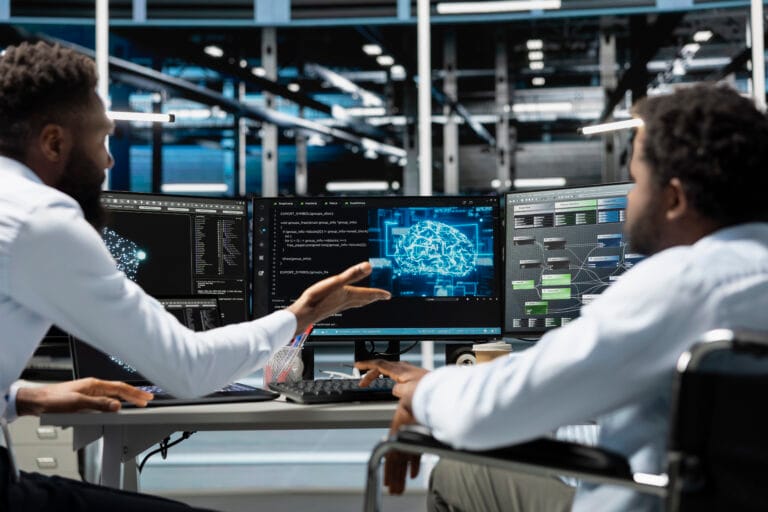 Man in wheelchair and colleague in data center running AI tools - NIHCF - NIHCF - NIHCF Two engineers collaborate at a workstation, monitoring three screens showing code and a holographic brain graphic in a tech office setting.