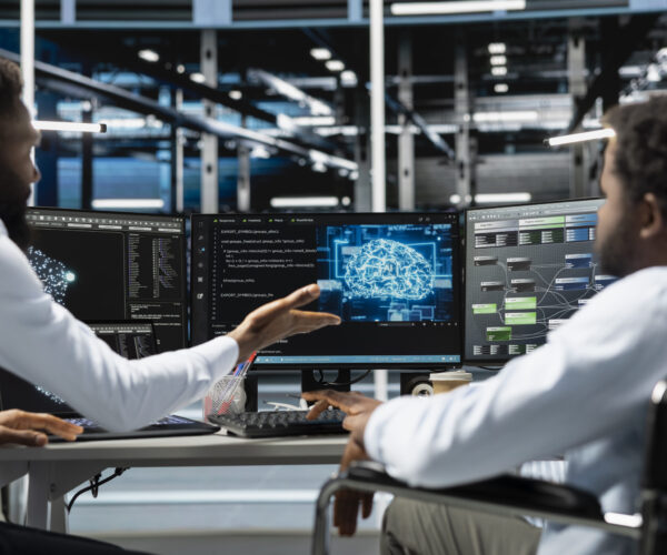 Two engineers collaborate at a workstation, monitoring three screens showing code and a holographic brain graphic in a tech office setting.