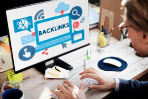 Person typing on a keyboard as a monitor displays a blue sketch with the word BACKLINKS, suggesting SEO work.