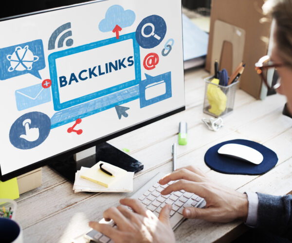 Person typing on a keyboard as a monitor displays a blue sketch with the word BACKLINKS, suggesting SEO work.